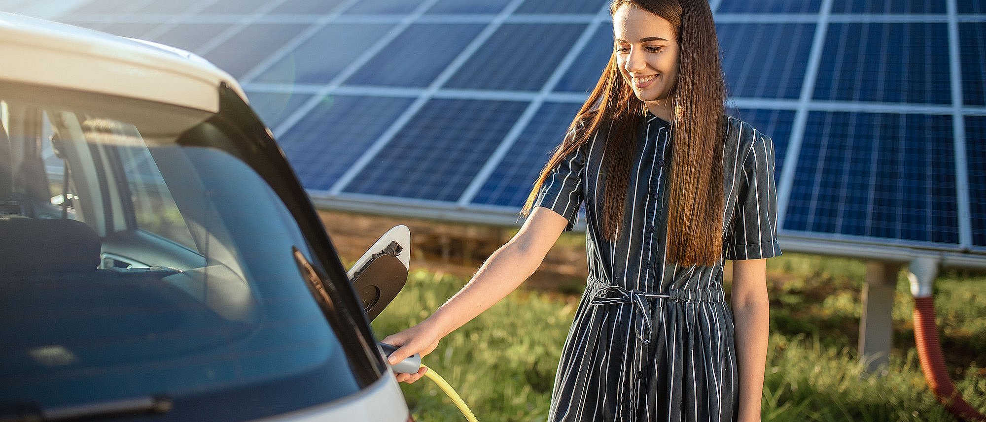 Lächelnde Frau lädt E-Auto und im Hintergrund ist eine große Photovoltaikanlage auf einer grünen Wiese